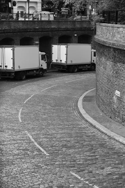 A black and white image showing two medium-sized white commercial vans parked on a cobblestone driveway near a brick wall and an arched underpass. The vans are positioned side by side, with the loading areas facing toward a curved section of the driveway. The scene is outdoors, with a metal railing and trees visible on the elevated area behind the wall. The driveway is clean and free of obstructions, indicating a space used for moving activities, such as home relocation or furniture transport. The surrounding environment suggests an urban or residential setting, with no visible moving personnel or equipment in the image. This setting illustrates the logistical aspect of house removals, with the vans ready for loading or unloading furniture, boxes, and packing materials, aligned with the services provided by Kennington Man and Van in their house removal operations.
