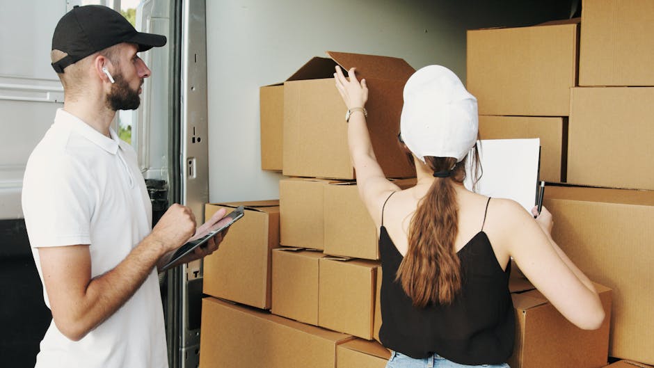 A man and a woman are working together next to a large vehicle used for house removals in an indoor loading area. The man, wearing a white polo shirt, a black cap, and wireless earbuds, is holding a tablet and observing the process. The woman, wearing a white protective cap and a black sleeveless top, is actively reaching up to a stack of neatly arranged cardboard boxes, some partially open, which are positioned on the floor and against the vehicle's interior wall. The boxes vary in size and are sealed with packing tape. The woman holds a clipboard in her other hand, indicating organisation during packing or loading. The environment is well-lit, with the vehicle's interior visible behind them, ready for furniture and packing materials to be loaded for a home relocation. This scene depicts the detailed packing and loading process associated with professional removals services, such as those provided by Kennington Man and Van, highlighting efficient furniture transport, packing materials, and careful handling during a house move.