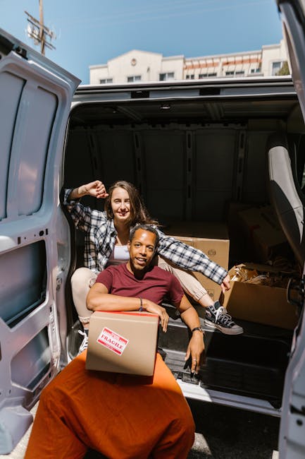 Two women engaged in home relocation activities with a van parked outside an urban building under clear skies. One woman, seated on the ground, holds a cardboard box marked 'fragile' and smiles at the camera, while the other woman stands behind her inside the open rear cargo area of a silver panel van, which is filled with several cardboard moving boxes, wrapping materials, and a small piece of furniture. The woman inside the van is wearing a checkered shirt and jeans, lifting a box with one hand and gesturing with the other. The scene captures the loading process during a house move, with some boxes arranged inside the vehicle and others being carried, indicating a professional packing and furniture transport operation by Kennington Man and Van in the context of estate and house removals.