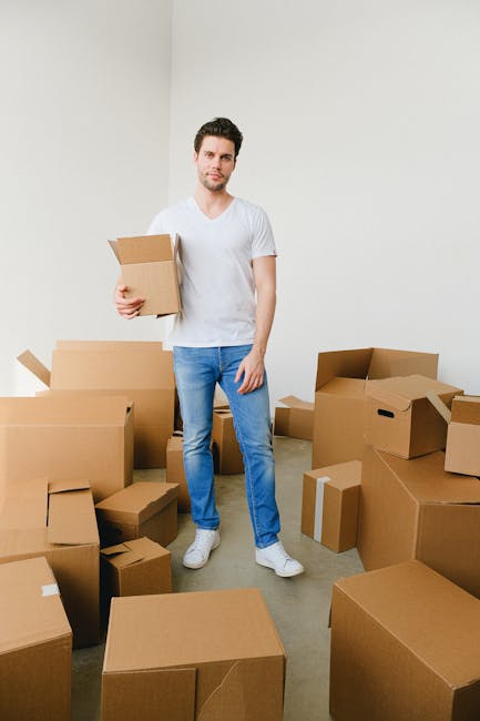A young man with short dark hair and a beard, dressed in a white t-shirt and blue jeans, is standing inside a room surrounded by numerous cardboard boxes of various sizes, some sealed with packing tape and others open. He is holding a medium-sized cardboard box in his right hand while looking at the camera with a neutral expression. The room has plain white walls and a light-colored carpeted floor. There are no other furnishings or items visible in the space. This scene illustrates a home relocation process, captured during packing or unpacking stages, with the boxes arranged around him. The environment suggests an ongoing house move, and the presence of multiple boxes indicates a significant household move, as handled by professional moving services such as Kennington Man and Van, referenced on the page about estate moves in Brandon Estate. The natural lighting in the room and the everyday setting support the depiction of a typical house removal or furniture transport process.
