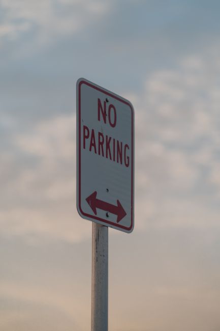 A tall rectangular white sign with a red border and bold red 'NO PARKING' text, positioned on a metal pole against a partly cloudy sky background. The sign features a red double-headed arrow at the bo