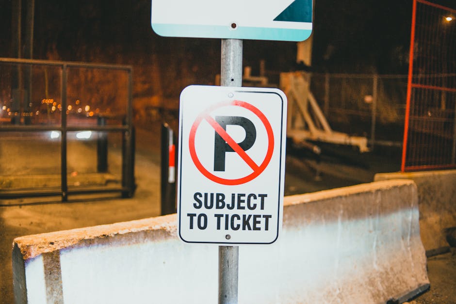 A tall rectangular white sign with a red border and bold red 'NO PARKING' text, positioned on a metal pole against a partly cloudy sky background. The sign features a red double-headed arrow at the bo