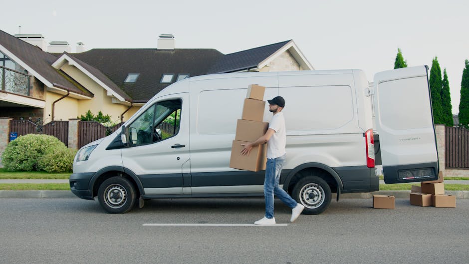 Two women are engaged in home relocation activities with a van parked outdoors in an urban setting, with multi-storey buildings in the background under a clear blue sky. One woman, wearing a plaid shi