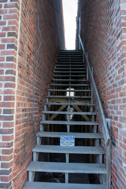 A narrow outdoor staircase made of unfinished wooden planks with horizontal treads, set between two brick walls that extend vertically on either side. The staircase features a simple wooden handrail on the right side, running parallel to the steps, and is elevated above the ground with supporting wooden beams visible beneath. A small notice sign at the base of the staircase reads, 'NOTICE EMPLOYEES ONLY BEYOND THIS POINT,' indicating restricted access. The setting appears to be part of a residential or commercial building's back or side alleyway, with natural daylight illuminating the scene from above and between the brick walls. The image reflects a typical obstacle encountered during house removals or furniture transport through tight, old staircases often found in urban properties, which complicates home relocation efforts for moving specialists like Kennington Man and Van working within properties with limited access.