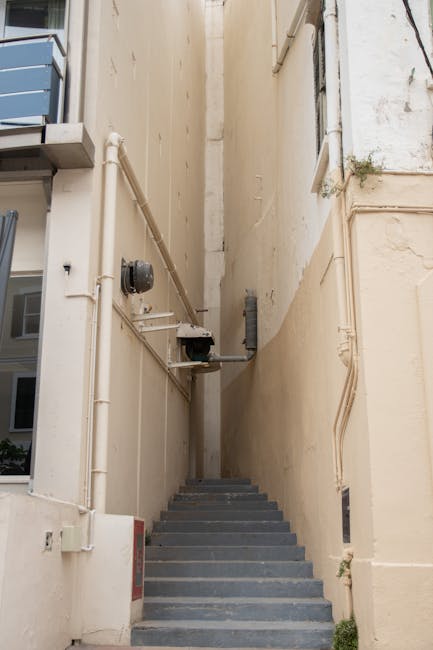 A narrow outdoor staircase made of unfinished wooden planks with horizontal treads, set between two brick walls that extend vertically on either side. The staircase features a simple wooden handrail on the right side, running parallel to the steps, and is elevated above the ground with supporting wooden beams visible beneath. A small notice sign at the base of the staircase reads, 'NOTICE EMPLOYEES ONLY BEYOND THIS POINT,' indicating restricted access. The setting appears to be part of a residential or commercial building's back or side alleyway, with natural daylight illuminating the scene from above and between the brick walls. The image reflects a typical obstacle encountered during house removals or furniture transport through tight, old staircases often found in urban properties, which complicates home relocation efforts for moving specialists like Kennington Man and Van working within properties with limited access.