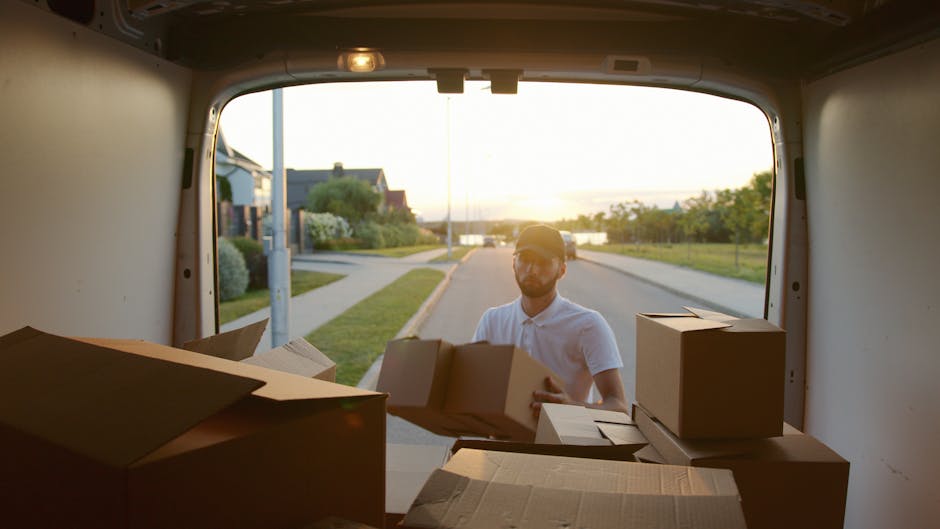 A man with a beard, wearing a white shirt and cap, is seen loading cardboard boxes into the back of a moving van parked on a suburban street during sunset. The van’s interior is visible, showing several sealed and unsealed cardboard cartons, some opened to reveal packing materials like bubble wrap and packing paper. The boxes are stacked on the vehicle’s floor, with some being lifted or carried by the man, indicating the loading process for a house relocation or furniture transport. Outside the van, a quiet residential area is visible with houses, well-maintained lawns, bushes, and a walking path, illuminated by warm sunset light. The surroundings include a lamppost and water in the distance, suggesting proximity to a river or canal. This scene reflects a professional moving service, such as Kennington Man and Van, engaged in packing and loading household items for a local move, emphasizing logistical aspects of house removals and furniture transport.