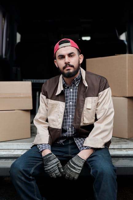 A young man with a dark beard, wearing a red baseball cap, a plaid shirt, a beige and brown jacket, and striped gloves, is seated on the edge of an open van trunk. Behind him are multiple closed cardboard boxes stacked inside the vehicle, which is parked on a paved area. The interior of the van is visible above his head, with a dark ceiling and rear seats. The scene depicts the loading process during a home relocation or furniture transport, with the man taking a break amidst the packing and moving activities associated with a professional removals service, such as Kennington Man and Van.