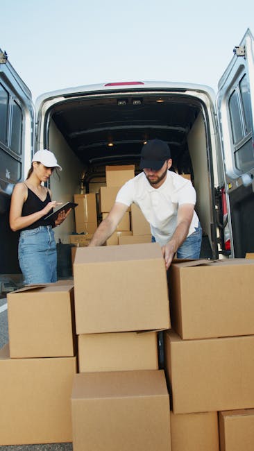 A man with a beard, wearing a white shirt and cap, is seen loading cardboard boxes into the back of a moving van parked on a suburban street during sunset. The van’s interior is visible, showing several sealed and unsealed cardboard cartons, some opened to reveal packing materials like bubble wrap and packing paper. The boxes are stacked on the vehicle’s floor, with some being lifted or carried by the man, indicating the loading process for a house relocation or furniture transport. Outside the van, a quiet residential area is visible with houses, well-maintained lawns, bushes, and a walking path, illuminated by warm sunset light. The surroundings include a lamppost and water in the distance, suggesting proximity to a river or canal. This scene reflects a professional moving service, such as Kennington Man and Van, engaged in packing and loading household items for a local move, emphasizing logistical aspects of house removals and furniture transport.