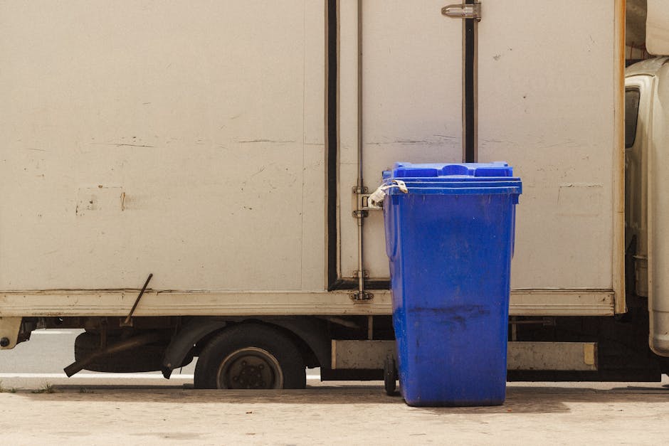 A large collection of overflowing waste and recycling bins placed on a paved sidewalk in front of a commercial building. The waste includes cardboard boxes, plastic bags, paper, and household items, s