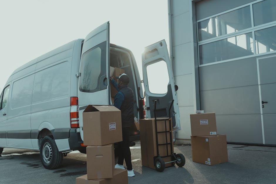 A worker from Kennington Man and Van is engaged in a home relocation process, loading cardboard boxes into the back of a large white moving van parked outside a modern building with glass windows. The worker, dressed in a dark uniform with a cap, is standing at the open rear doors of the van, placing a medium-sized cardboard box inside. Several other packed boxes, some with labels, are positioned on the pavement near the van, along with a black metal trolley with wheels and lifting straps nearby, indicating the packing and loading process. The van is situated adjacent to the building's entrance, which features a large glass door and window panels, with sunlight creating a bright, clear atmosphere. The scene captures the logistics involved in furniture transport and house removals, emphasizing the careful handling of packed items during a timely moving service, consistent with the focus on last-minute van hire in SE11, as offered by Kennington Man and Van.