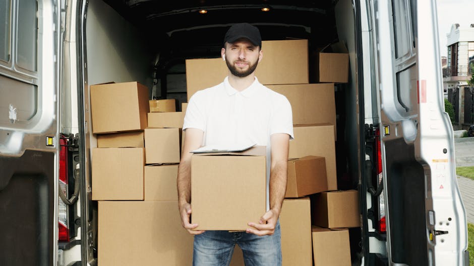 A man with a beard and wearing a black cap, white polo shirt, and jeans is standing at the rear of a white moving van, holding a cardboard box with both hands. The van's interior is filled with various cardboard boxes of different sizes, some stacked on top of each other, ready for a home relocation or furniture transport. The boxes are sealed with packing tape, and some are supported with plastic wrap to protect fragile contents. The van is parked outside a residential area, visible by the pavement and nearby houses. Inside the van, additional boxes are arranged on the floor and against the walls, with a few larger pieces of furniture or storage units partially visible in the background. The scene captures the loading process of packing and moving belongings during a house removal, with professional services from Kennington Man and Van evident in the organized setup for efficient house moving or last-minute relocation in SE11.