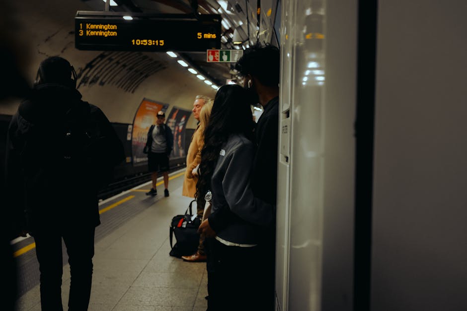 Inside an underground train station, several passengers are waiting on the platform for their train. The platform features a dark ceiling with built-in lighting, and an electronic display board showing train departure times and destinations, including Kennington. Passengers are seen standing near the edge, some carrying backpacks or luggage, with one person partially visible leaning against the platform wall. The background shows the train track area and another train approaching or stopped at the station. The scene captures the typical environment of a city travel hub, where people prepare for home relocation or moving logistics, and [COMPANY_NAME], such as Kennington Man and Van, supports the transport and packing process involved in residential removals. The image highlights the importance of efficient furniture transport and packing during busy commuting hours, with materials like fabric and cardboard visible near luggage and seating areas, while the overall environment remains clean and well-lit for safety and accessibility.