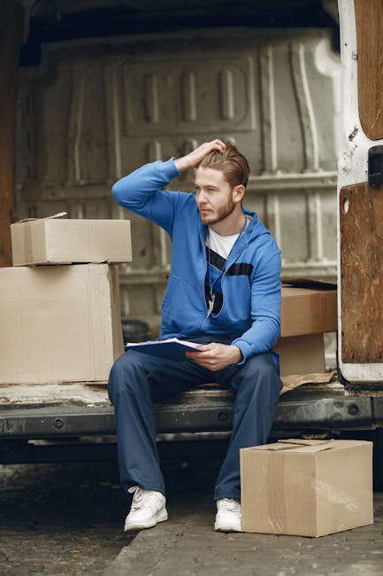 A young man with light brown hair, dressed in a blue hoodie with dark blue accents, sitting on the edge of a vehicle's loading area during a home relocation process. He is holding a clipboard in his left hand and appears to be contemplating, with his right hand resting on his head. Surrounding him are several cardboard boxes of various sizes, some placed directly on the ground and others stacked beside him, ready for packing or unpacking. The interior of the vehicle is visible behind him, showing a metal panel with a slightly textured surface and a white interior wall. The scene suggests a moving or packing scenario, with protective materials like cardboard boxes, indicating careful handling of belongings. The vehicle is parked outside, and the lighting appears natural, typical of daytime. The presence of the boxes and the vehicle emphasizes the process of furniture transport, packing, and loading involved in house removals, which is part of the services offered by Kennington Man and Van.