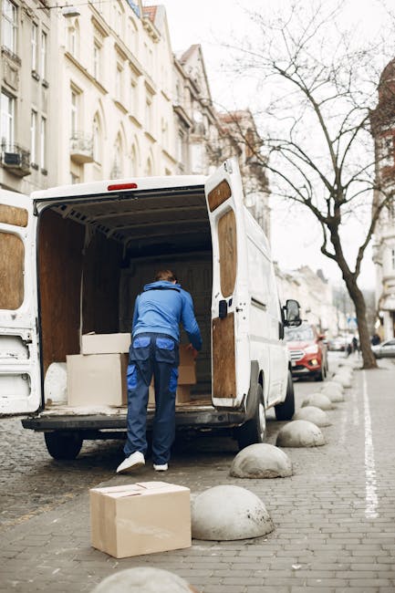 A man dressed in a blue hoodie and dark trousers is loading large white cardboard boxes into the back of a white commercial van parked on a cobblestone street. The van's rear doors are open, revealing an empty interior with wood paneling. The street features a line of parked cars and several large, smooth concrete street bollards along the pavement. In the background, there are multi-storey residential buildings with ornate facades and balconies, alongside leafless trees indicating a colder season. The scene captures a home relocation or furniture transport process, with the man actively involved in packing or unloading items, supported by the van and packing materials. The natural daylight provides neutral lighting, emphasizing a professional and organized moving operation, aligning with the removals services provided by Kennington Man and Van.