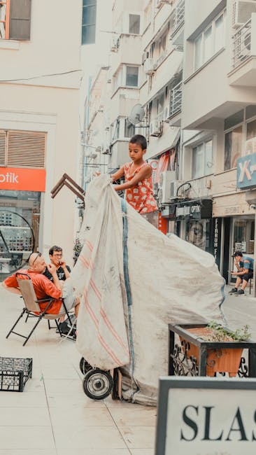 A young boy standing on a hand truck or sack trolley outdoors on a busy urban street, loading or unloading large fabric or plastic shipments. Surrounding him are colleagues or passersby, some seated in outdoor chairs and others walking past. The background features multi-storey residential or commercial buildings with balconies, satellite dishes, and shop fronts including a pharmacy, indicating a lively commercial area. The scene appears to involve a packing or moving process, with the boy lifting or positioning the material on the trolley, which is commonly used during house removals. The environment is bright with natural daylight, and the pavement surface is smooth, consistent with an active city street. This setting supports themes of logistics, furniture transport, and home relocation services, and is naturally aligned with [COMPANY_NAME]'s offerings described on the page for small flat removals near Kennington Station.