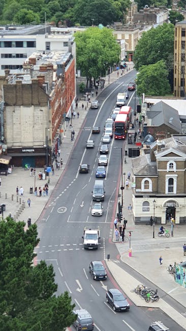 An aerial view of a busy urban street near Kennington Station showing multiple vehicles including cars, vans, and double-decker buses traveling along the road, with a mix of parked and moving cars on the street. On the right side of the image, there is a small white building with a pitched roof, adjacent to a pavement where pedestrians are walking. Several bicycles are parked near the building, and a few pedestrians are crossing the street at designated crosswalks. Large, leafy green trees line both sides of the street, partially obscuring some buildings, including a red brick structure on the left and a modern white building in the background. The scene appears to be during daytime with natural light illuminating the area, capturing the typical environment of London’s residential and commercial districts, relevant to house removals and moving logistics as undertaken by companies like Kennington Man and Van.